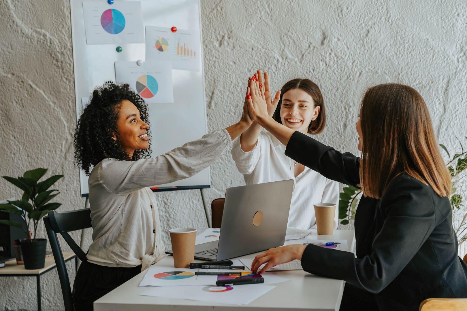 Tres mujeres profesionales celebran un logro con un "high five" en una reunión de trabajo, frente a gráficos y ordenador portátil.