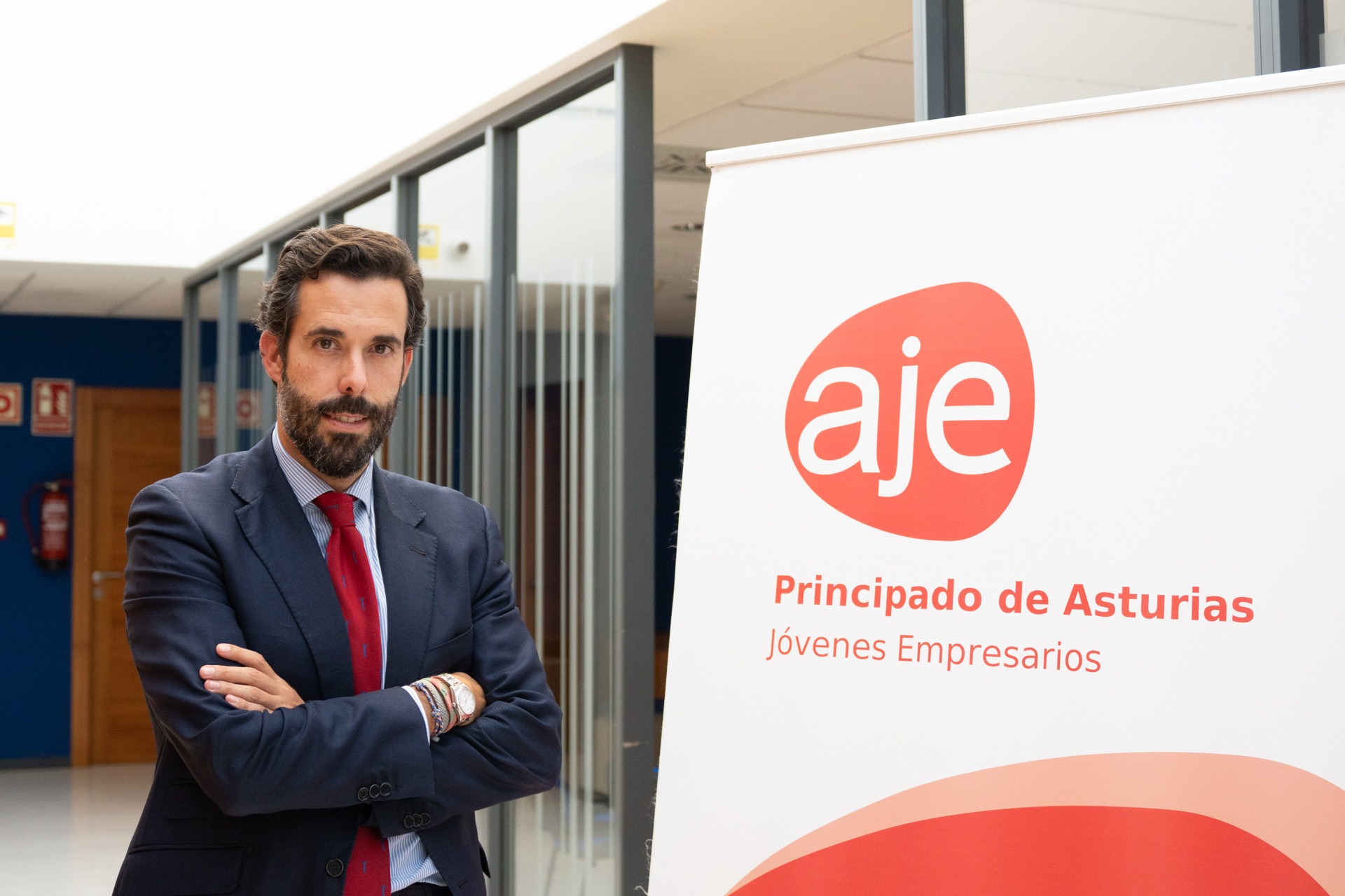 Hombre con traje y corbata roja posando junto a un cartel de AJE Principado de Asturias, Jóvenes Empresarios.